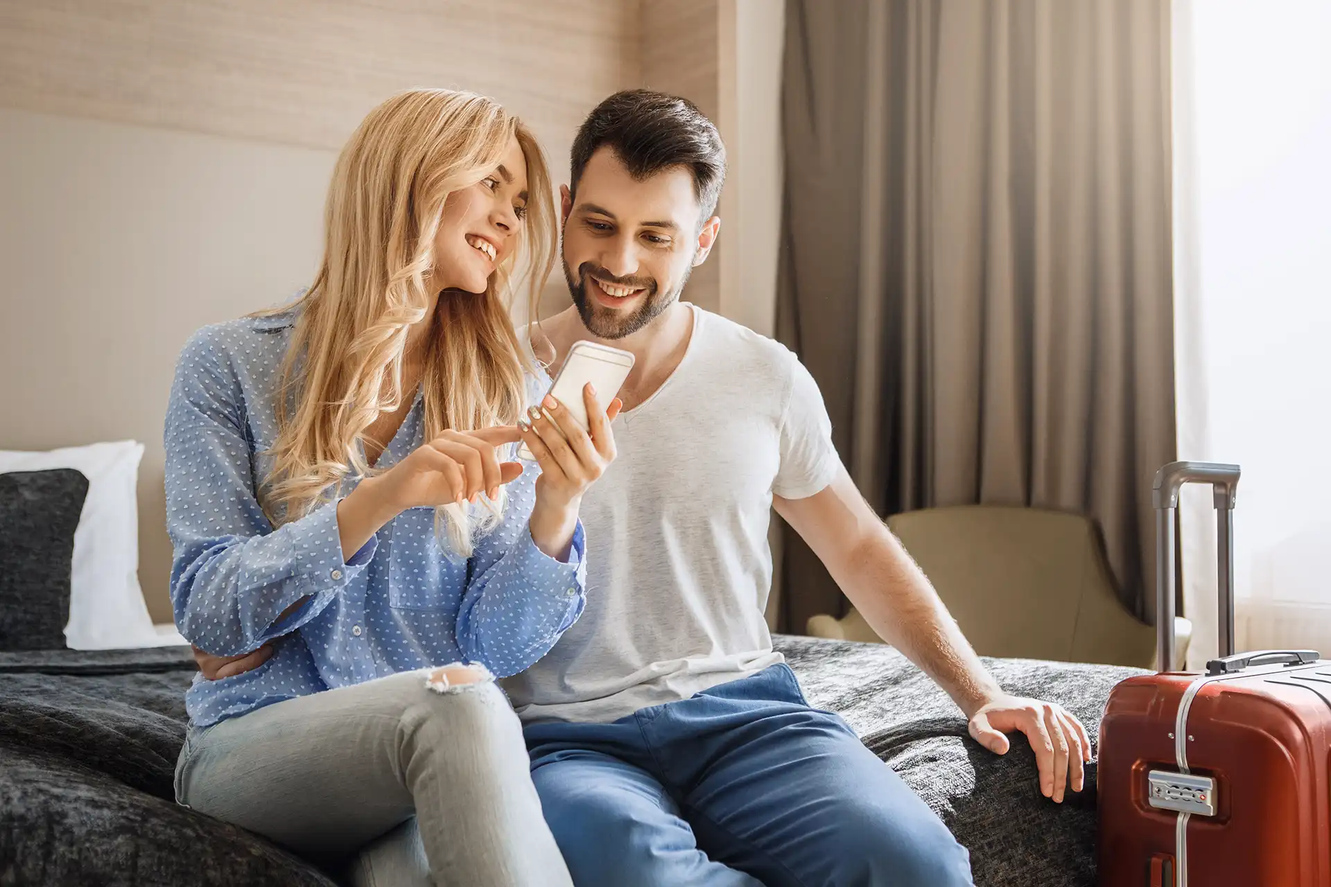 Young Couple Look At Phone While Sitting On Hotel Bed.